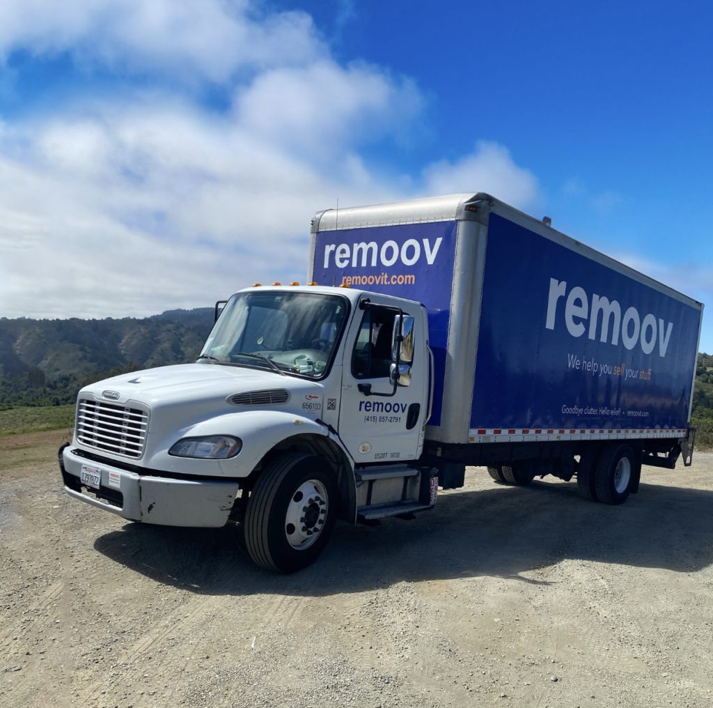 Image of a blue and white moving truck with the Remoov logo on the side. Truck is parked in a parking lot in front of a blue sky.